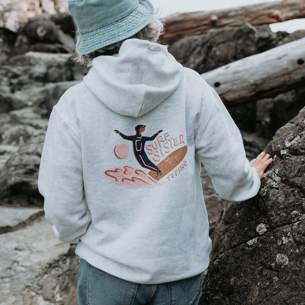 A person standing outdoors on a rocky shore, shown from the back wearing a light heather gray hoodie with a colorful back graphic of a surfer and the words “Surf Sister” 