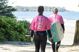 Two people in pink rash guards walking on a beach with surfboards.
