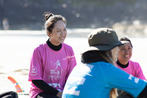 Two women in pink and blue rash guards sitting by a body of water, smiling during a surf lesson. 