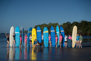 A group of surfers pose with a lineup of colorful surfboards on a beach. The surfboards are in various colors including blue, yellow, white, and beige, with the group wearing swimwear.