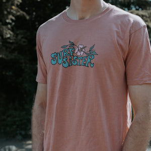 A man wearing a dusty pink t-shirt with a retro-style 'SURF SISTER' graphic in teal and white on the chest, set against a blurred outdoor background.