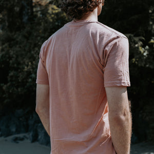 A man viewed from behind wearing a dusty rose  t-shirt, standing outdoors against a dark, blurred background of trees.