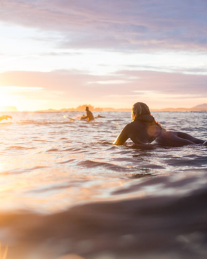 Person laying on surfboard in the ocean with a sunset or sunrise in the background