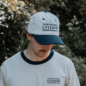 A man wearing a cream and navy blue corduroy baseball cap with 'TOFINO SURF LIFESAVING CLUB' printed on the front, paired with a matching white t-shirt with a dark navy collar, set against a blurred outdoor background of green foliage.