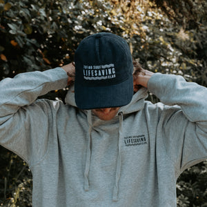 A person outdoors wearing a dark navy corduroy cap with white 'TOFINO SURF LIFESAVING CLUB' text and a light heather gray hoodie with matching text on the chest. The background is a blurred natural outdoor setting.