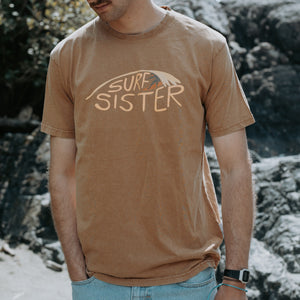 A man wearing a brown t-shirt with a 'SURF SISTER' graphic, standing outdoors in front of a rocky background.