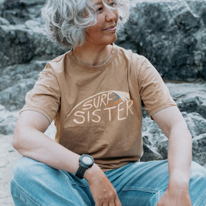 A woman with curly gray hair sits outdoors among rocks, wearing a brown graphic T-shirt that reads 'SURF A SISTER' in white and orange, paired with blue jeans.