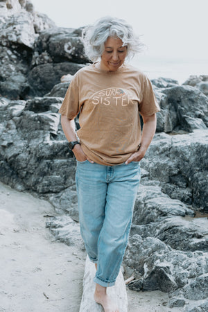 A woman with grey hair walking on a rocky beach wearing a brow t-shirt with 'SURF SISTER' printed across the chest, paired with light blue jeans.