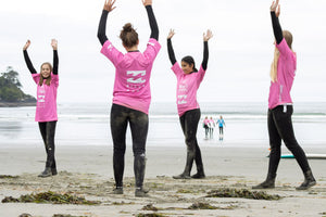 Four women in wetsuits and pink rash guards standing on a beach with arms raised.