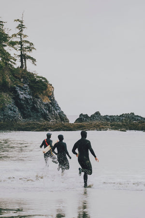 Three people in wetsuits running into the ocean with trees and rocks in the background.