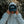 A person outdoors wearing a light gray hoodie and a dark navy cap with 'TOFINO SURF LIFE SAVINGS CLUB' printed on the front. The person is holding the hood of the sweatshirt up over their face, with blurred foliage in the background.