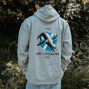 A man viewed from behind wearing a light heather gray hoodie with a large back graphic showing a seal over a blue-and-white shield and the text 'TOFINO SURF LIFESAVING CLUB'. The background is an outdoor setting with dark green foliage.