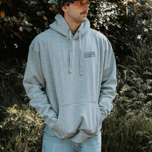 A man wearing a light gray heathered hoodie with a small black text graphic on the left chest, standing outdoors in front of a leafy background.
