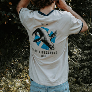 A man outdoors wearing a white ringer T-shirt with dark navy trim and a large back graphic featuring a seal over a blue shield, with the text 'TOFINO SURF LIFESAVING CLUB'.
