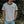 A man wearing a light beige ringer t-shirt with dark navy blue trim on the collar and sleeve cuffs, featuring a small black graphic print on the left chest. He is standing outdoors in front of a blurred green foliage background.