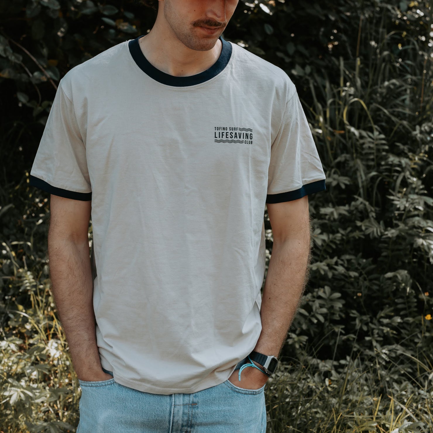 A man wearing a light beige ringer t-shirt with dark navy blue trim on the collar and sleeve cuffs, featuring a small black graphic print on the left chest. He is standing outdoors in front of a blurred green foliage background.
