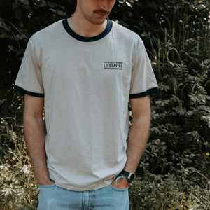 A man wearing a light beige ringer t-shirt with dark navy blue trim on the collar and sleeve cuffs, featuring a small black graphic print on the left chest. He is standing outdoors in front of a blurred green foliage background.