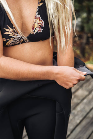 Person wearing a black floral bikini top, pulling on a black wetsuit outdoors.