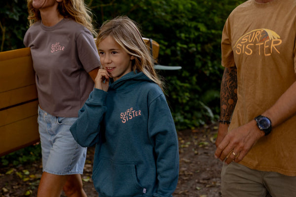 A young girl wearing a teal blue 'Surf Sister' hoodie stands outdoors in a wooded setting, flanked by two adults wearing graphic t-shirts. The girl's hoodie features the 'Surf Sister' brand name in a light pink script on the left chest.