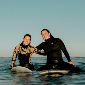 Two women in wetsuits sitting on surfboards in the water with a clear sky and smiling. 