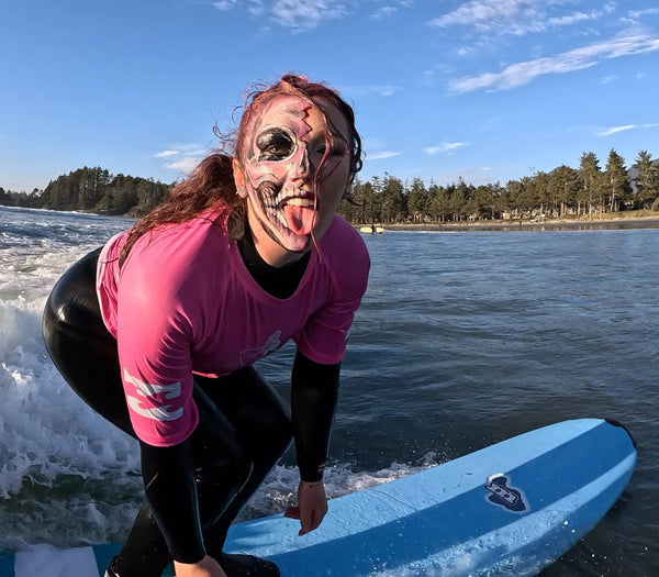 A woman in a pink and black wetsuit crouches on a blue surfboard in the water, wearing a skull face mask and sticking out her tongue. A beach and trees are visible in the background under a blue sky.