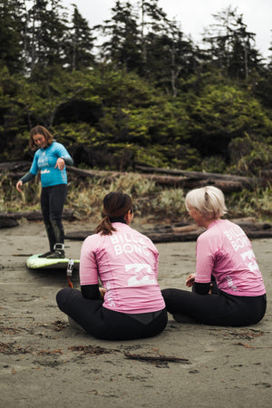 Three women on a beach with one standing on a surfboard and two sitting, all wearing pink rash guards