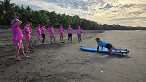 Group of people standing on the beach  in pink rash guards, watching a surf instructor demonstrate how to get on a surfboard 