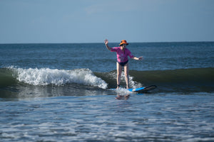 A ladystands on a surfboard in the ocean, wearing a bright pink long-sleeve top, black swim trunks, and an orange hat, with a small wave breaking behind them under a clear blue sky.