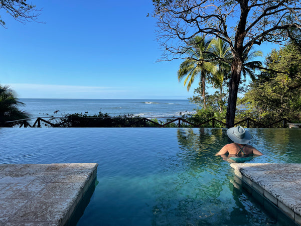 A woman wearing a wide-brimmed sun hat sits in an infinity pool overlooking a tropical ocean scene with palm trees and a clear blue sky.