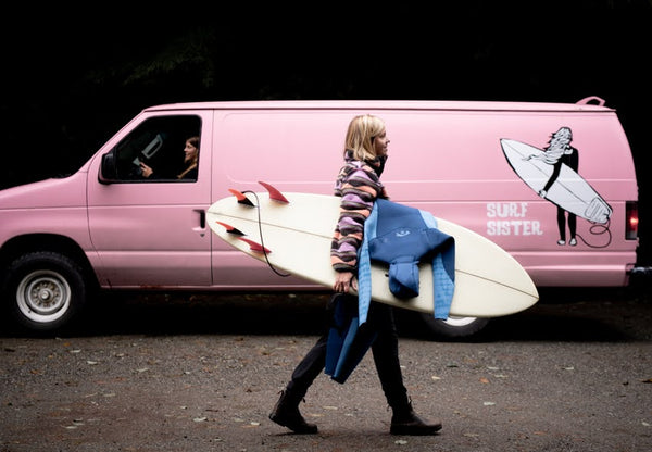 Woman walking infront of a pink van holding a surfboard 