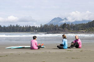 Three people sitting on a beach with surfboards, surrounded by trees and mountains.