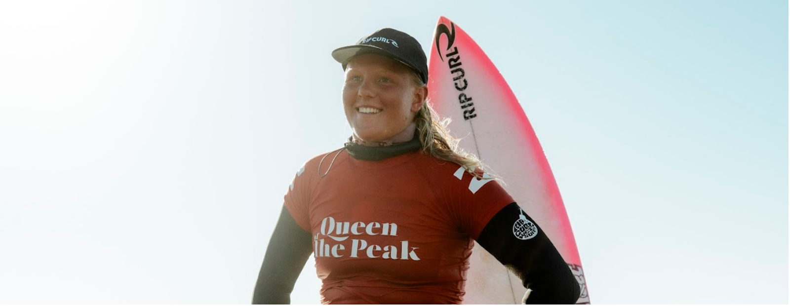Woman holding a surfboard with 'Rip Curl' branding, wearing a red rash guard with 'Queen of the Peak' text.