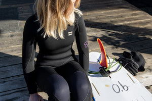 Person in a wetsuit sitting on a wooden dock with a surfboard and fins beside her.
