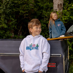 A boy wearing a light gray hoodie with a colorful 'Surf Sister' graphic stands in front of a dark truck, with a girl in a blue hoodie in the background among green trees.