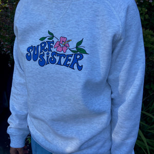 A person wearing a light heather gray crewneck sweatshirt with a blue 'SURF SISTER' graphic and a pink hibiscus flower on the chest, paired with blue jeans outdoors.
