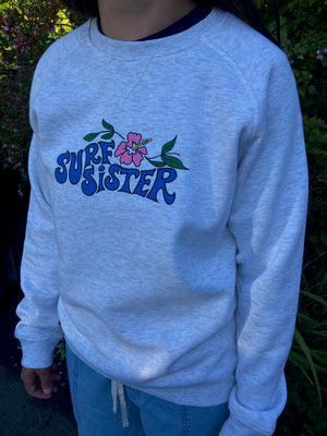 A person wearing a light heather gray crewneck sweatshirt with a blue 'SURF SISTER' graphic and a pink hibiscus flower on the chest, paired with blue jeans outdoors.