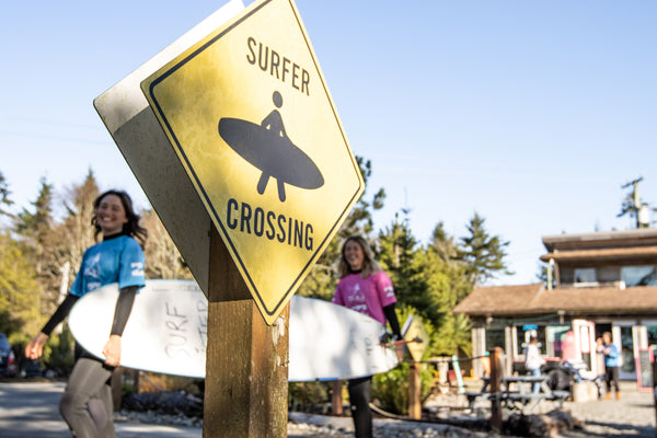 Surfer crossing sign with people carrying surfboards across a road with a surf shop in the background