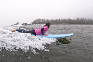 Woman laying on her stomach, surfing on a wave in a wetsuit with a pink rash guard and blue board.