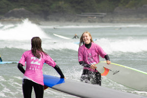 Two teenage girls in pink rash guards and wetsuits holding surfboards in the ocean
