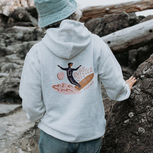 A person standing outdoors on a rocky shore, shown from the back wearing a light heather gray hoodie with a colorful back graphic of a surfer and the words “Surf Sister” 