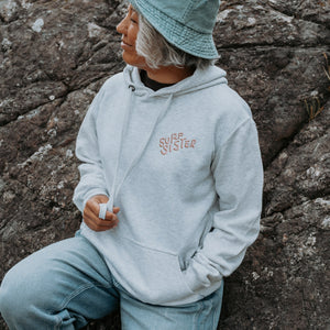 A lady sitting outdoors against a rocky surface wearing a light gray hoodie with 'SURF SISTER' logo on the chest. 