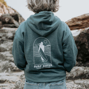 A person viewed from behind wearing a blue hoodie with a large white surf graphic and the text 'SURF SISTER, standing outdoors on rocky terrain.