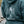 A close-up of a person wearing a blue hoodie outdoors. The back of the hoodie features a large white graphic of a surfer inside an arch with the text 'SURF SISTER' and 'TOFINO BC, CANADA'.