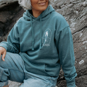 A lady wearing a blue pullover hoodie with a white surfer graphic on the chest and light blue jeans, seated outdoors against a rocky background.