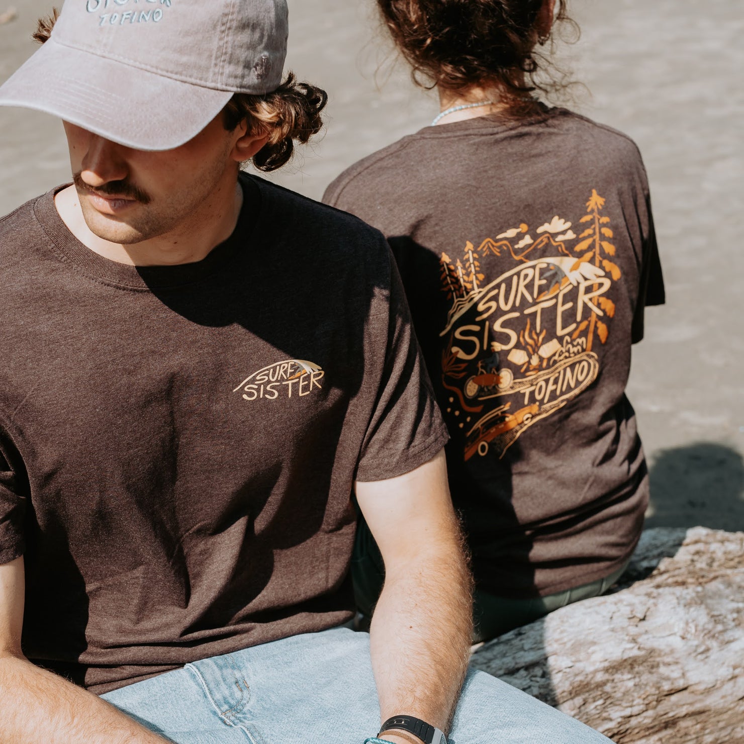Two people sitting on a log wearing the same brown t-shirt. The guy is facing the camera and the girl has her back. 