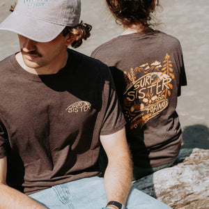 Two people sitting on a log wearing the same brown t-shirt. The guy is facing the camera and the girl has her back. 