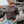 A woman with short gray hair wearing a dark brown t-shirt with a gold SURFER SISTER' logo on the left chest, paired with light blue jeans, leaning against a large piece of driftwood on a rocky beach.