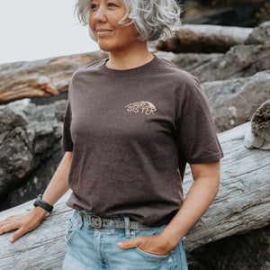 A woman with short gray hair wearing a dark brown t-shirt with a gold SURFER SISTER' logo on the left chest, paired with light blue jeans, leaning against a large piece of driftwood on a rocky beach.