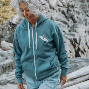 A woman with short gray hair wearing a blue zip-up hoodie with a white zipper and a small surfboard graphic on the chest, paired with light blue jeans, walking outdoors against a rocky background.