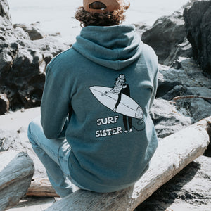 A man sitting on a log on a rocky beach, viewed from behind, wearing a blue hoodie with a white graphic of a surfer and the text 'SURF SISTER' on the back.